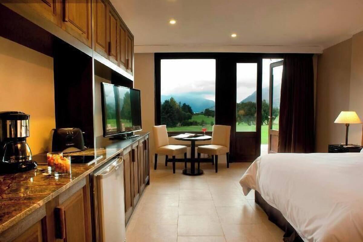 Habitación de hotel con cama, televisión, mesa de comedor con sillas, cafetera y vistas al exterior.