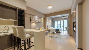 A modern hotel room with a kitchenette, a dining area, a living room with a sofa and a TV, and a large window.