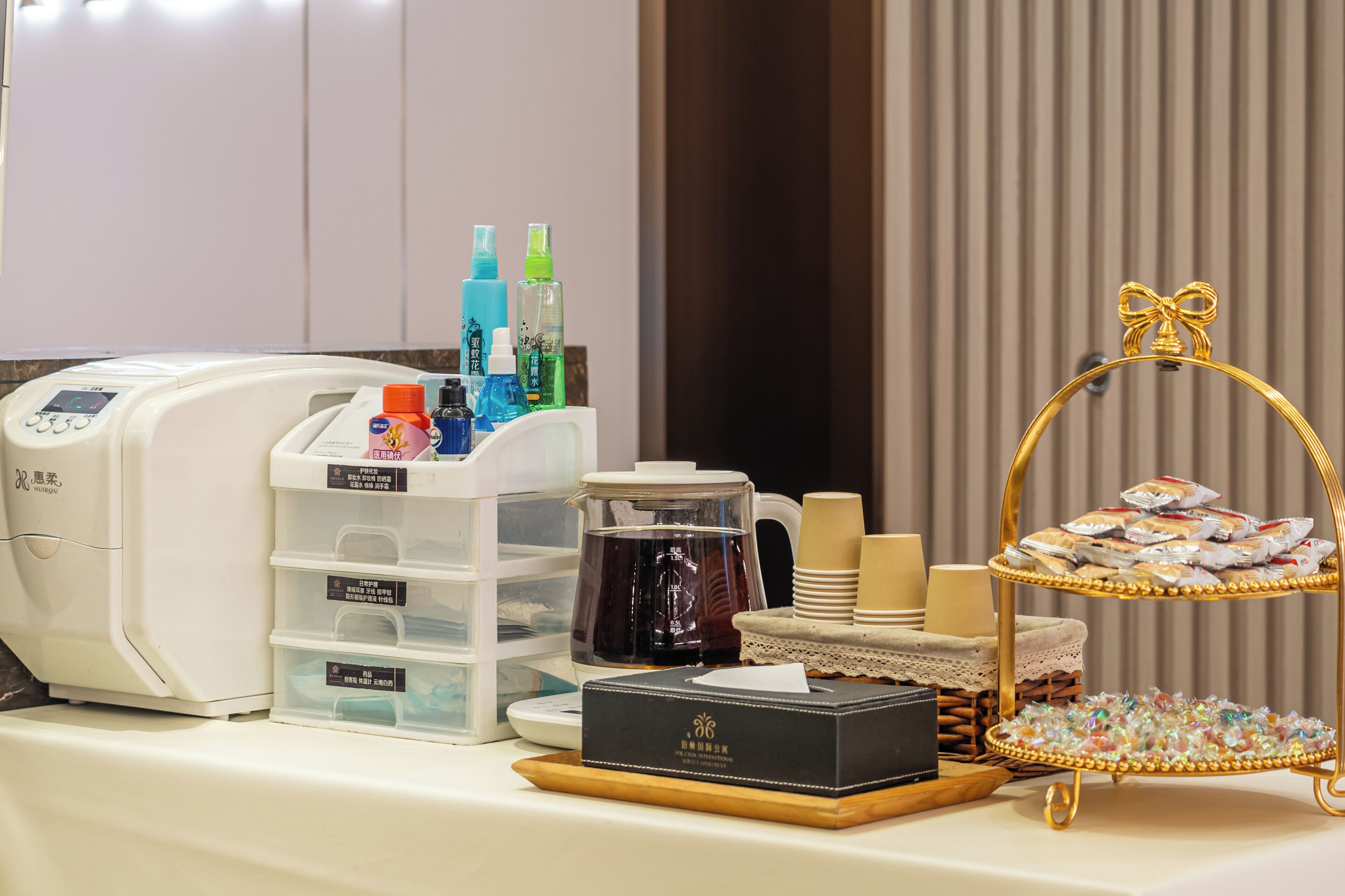 A table with a coffee maker, storage bins, a tea kettle, and a tray with snacks and a box.