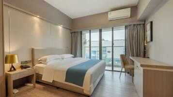 A hotel room with a bed, bedside table, lamp, desk, chair, and large windows.