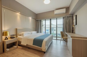 Una habitación de hotel con cama, mesita de noche, lámpara, escritorio, silla y ventanas amplias.