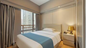 A hotel room with a bed, a nightstand, a lamp, and a view of tall buildings through the window.
