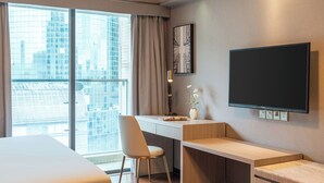 Una habitación de hotel moderna con una cama grande, un escritorio con una silla, un televisor de pantalla plana montado en la pared y un ventanal con vistas a la ciudad.