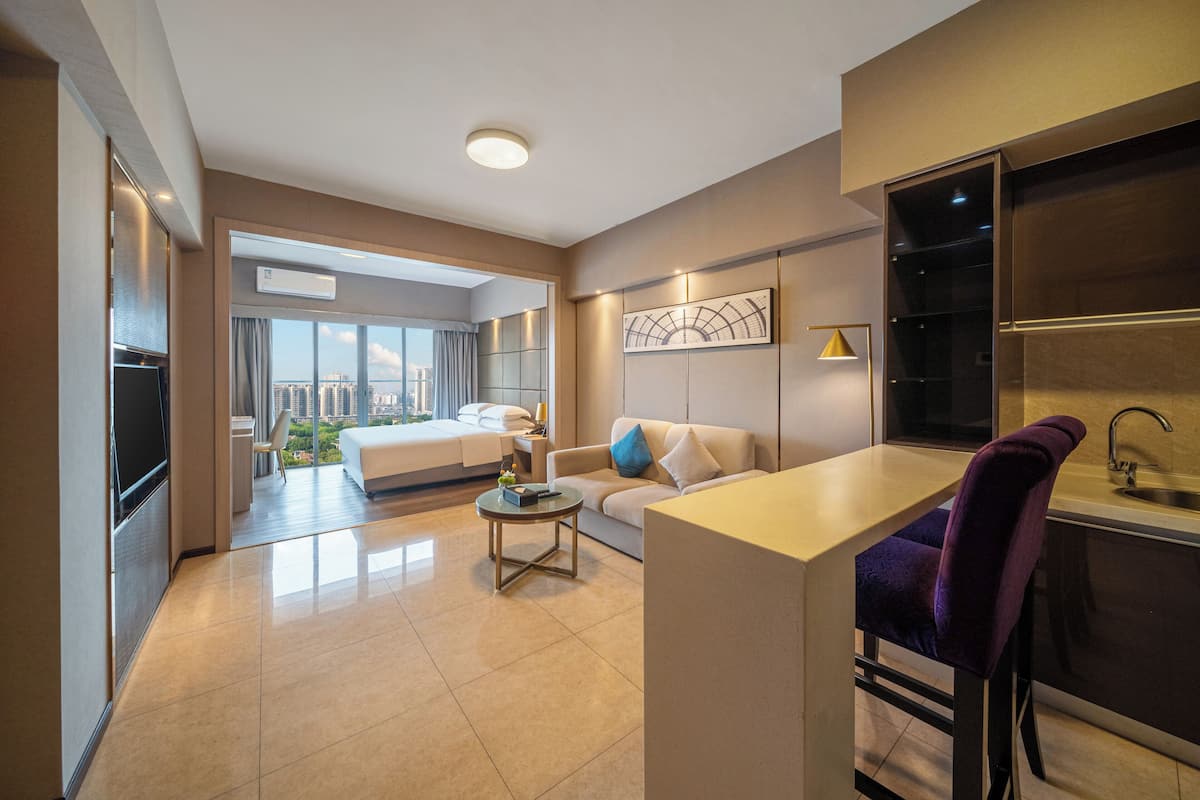 A modern hotel room with a large bed, a sofa, a dining area, and a kitchen.
