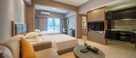 A modern hotel room with a sofa, a small round table, a bed with white bedding, a desk, and a kitchenette.