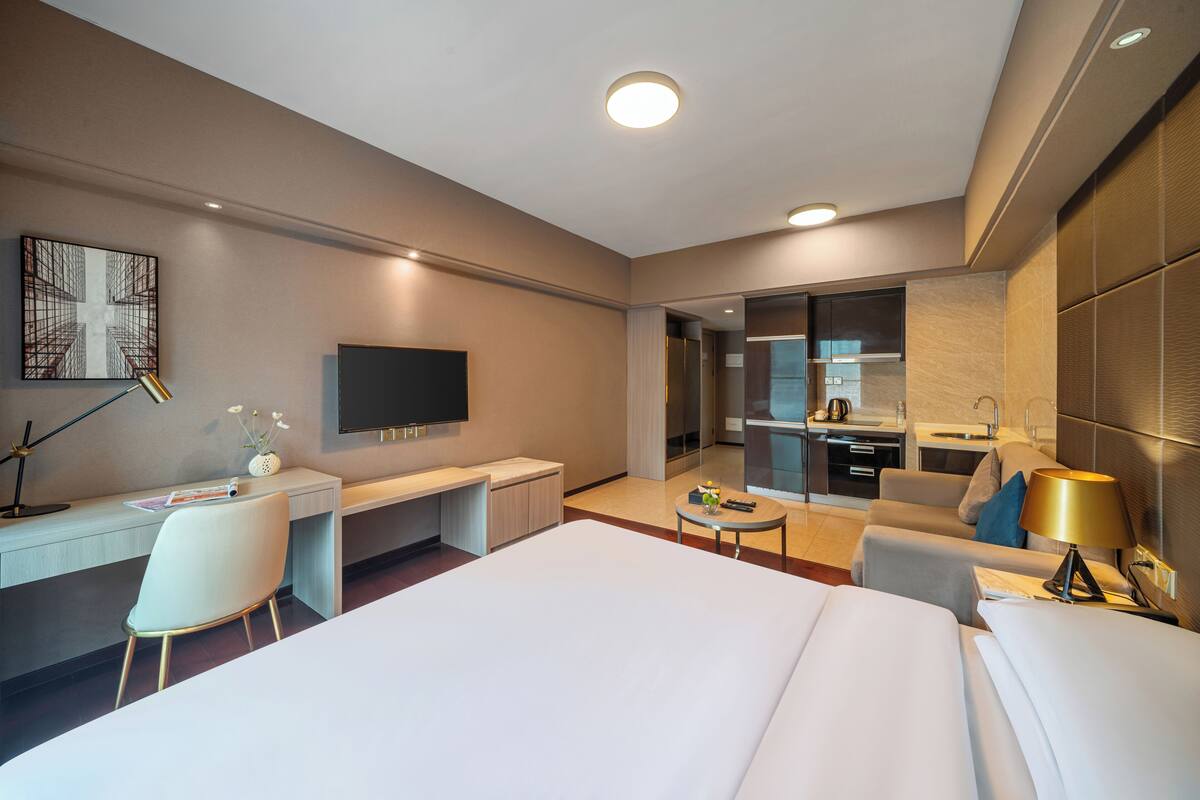 A modern hotel room with a large bed, a desk with a chair, a television, a small table with a lamp, and a kitchenette area.