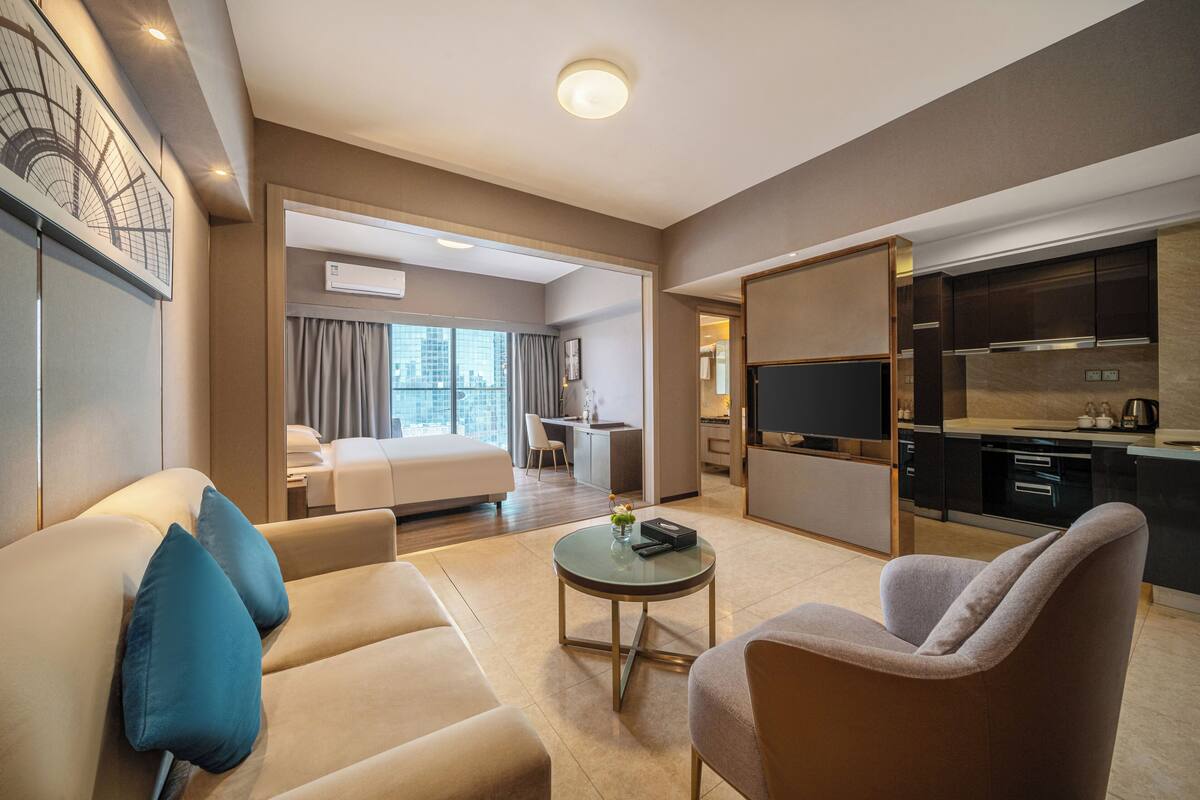 A modern hotel room with a living area, a bedroom, a kitchenette, and a large window offering a city view.