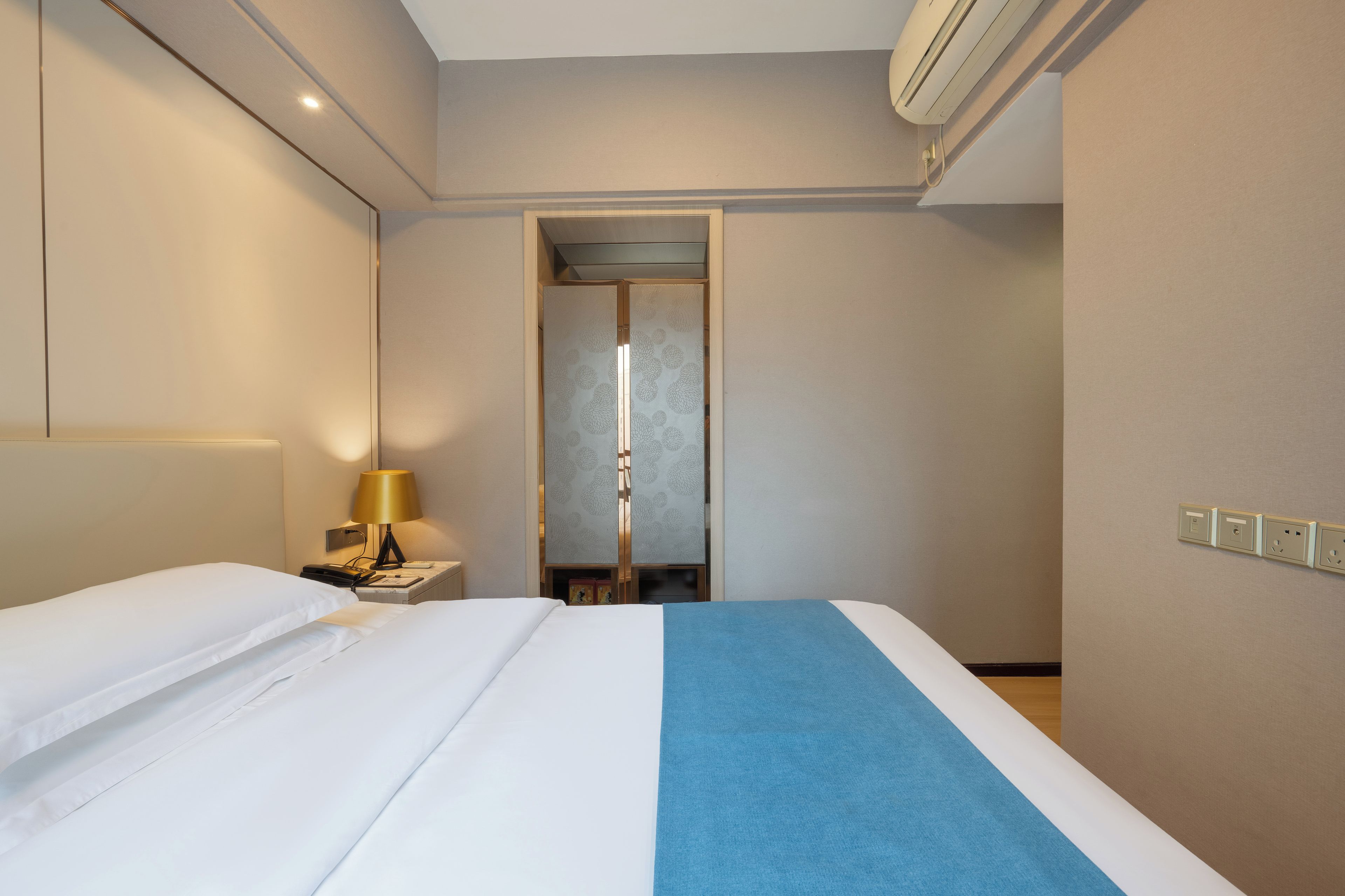 A hotel room with a bed, bedside table, lamp, and an air conditioning unit.