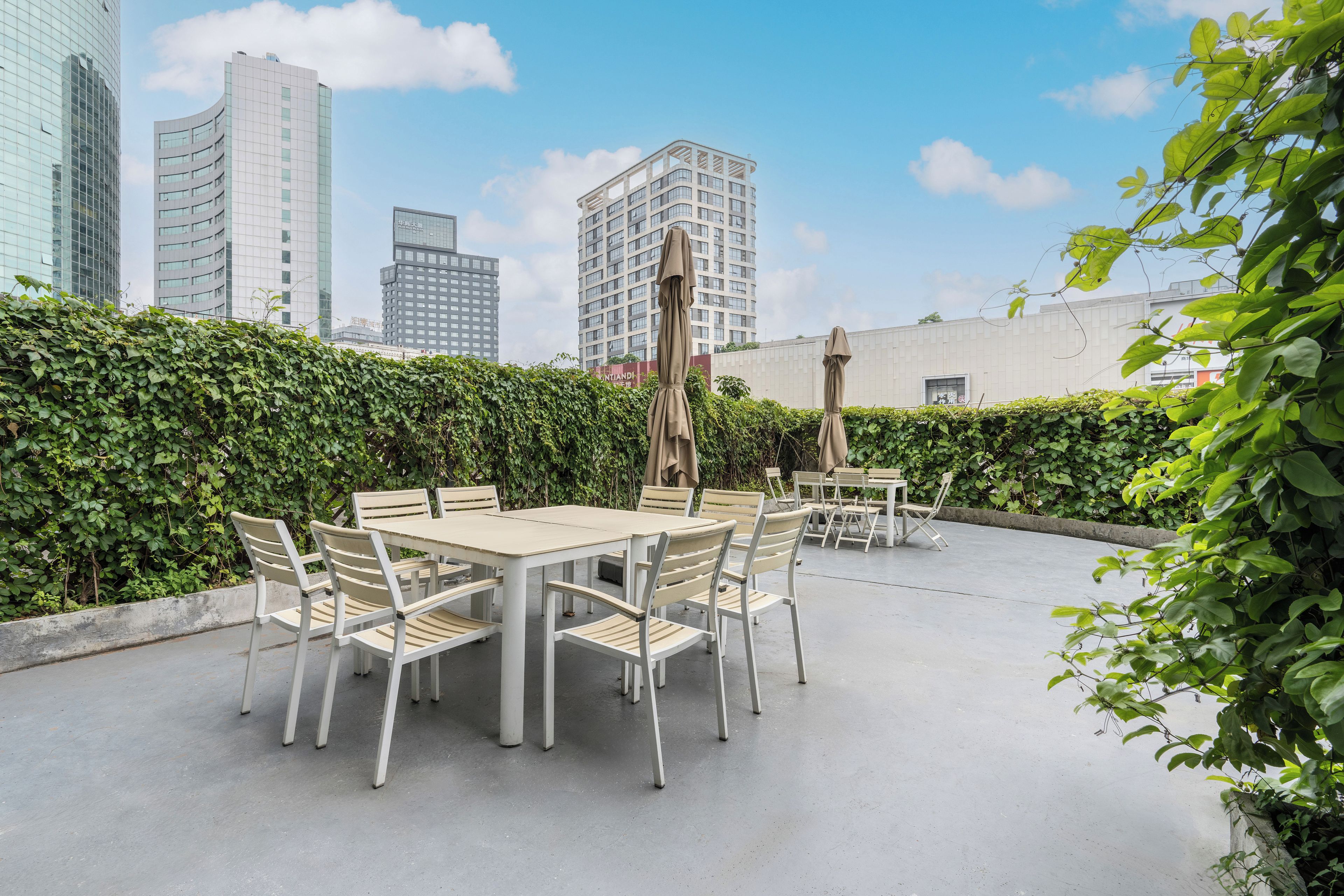An outdoor patio with a table and chairs, surrounded by greenery and overlooking a cityscape with modern buildings.