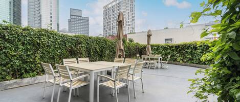 Een terras buiten met een tafel en stoelen, omgeven door groen en met uitzicht op een stadsgezicht met moderne gebouwen.
