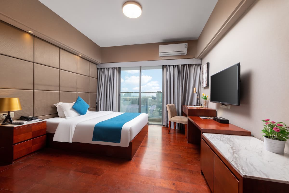 A hotel room with a large bed, a desk, a TV, and a balcony with a city view.