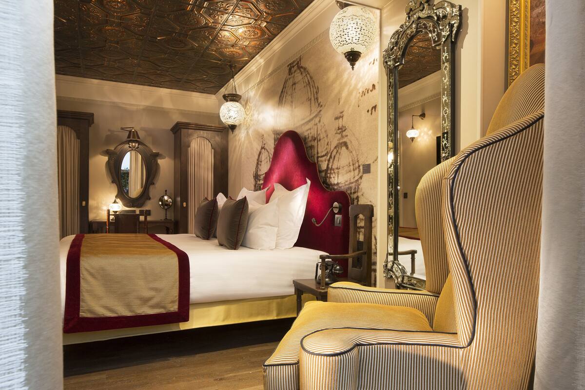 A hotel room with a large bed, a red headboard, a striped armchair, and ornate wall decorations.
