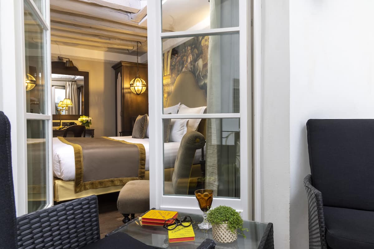 A hotel room with a bed, a nightstand, a lamp, a window, and a view of another room.