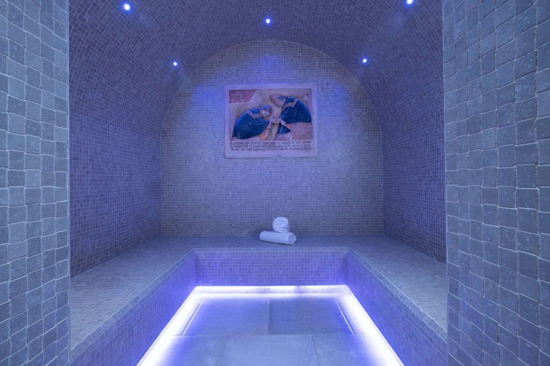Une salle de spa avec un sol et des murs carrelés, une affiche accrochée au mur du fond et un petit banc.