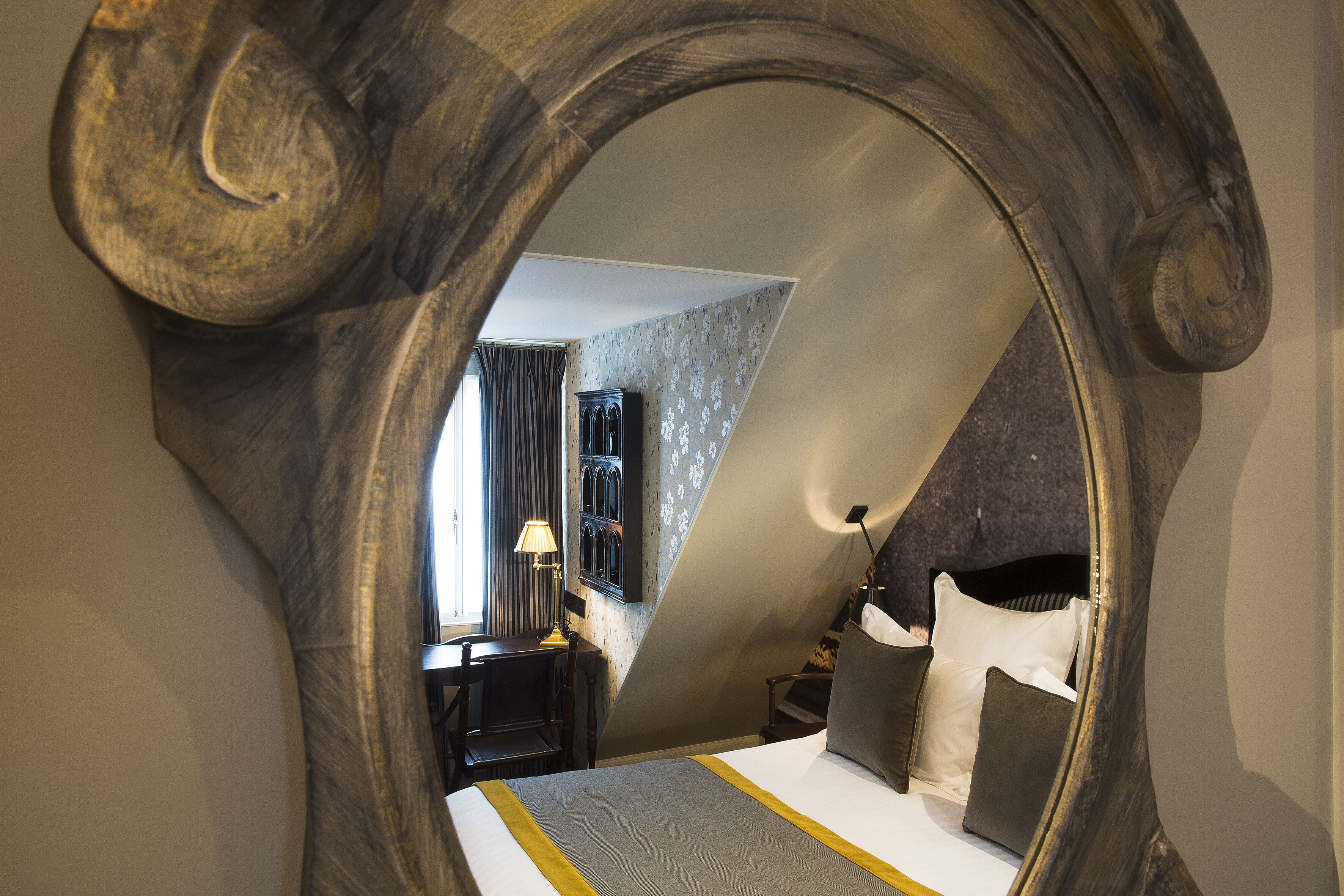 A bedroom with a large mirror reflecting the room, a bed with pillows, a nightstand, and a lamp.