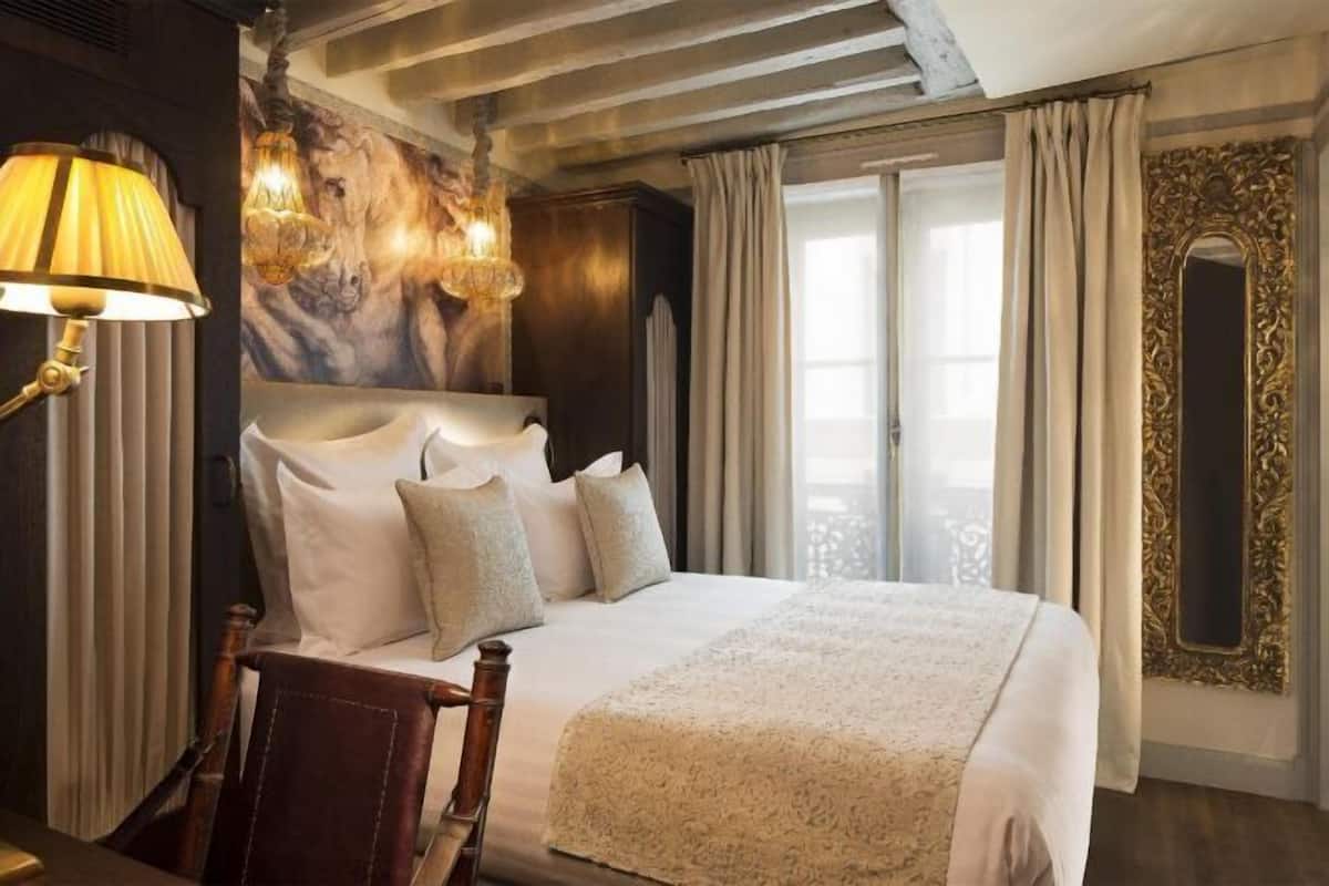 A hotel room with a large bed, a wooden chair, a lamp, a chandelier, and a large mirror.