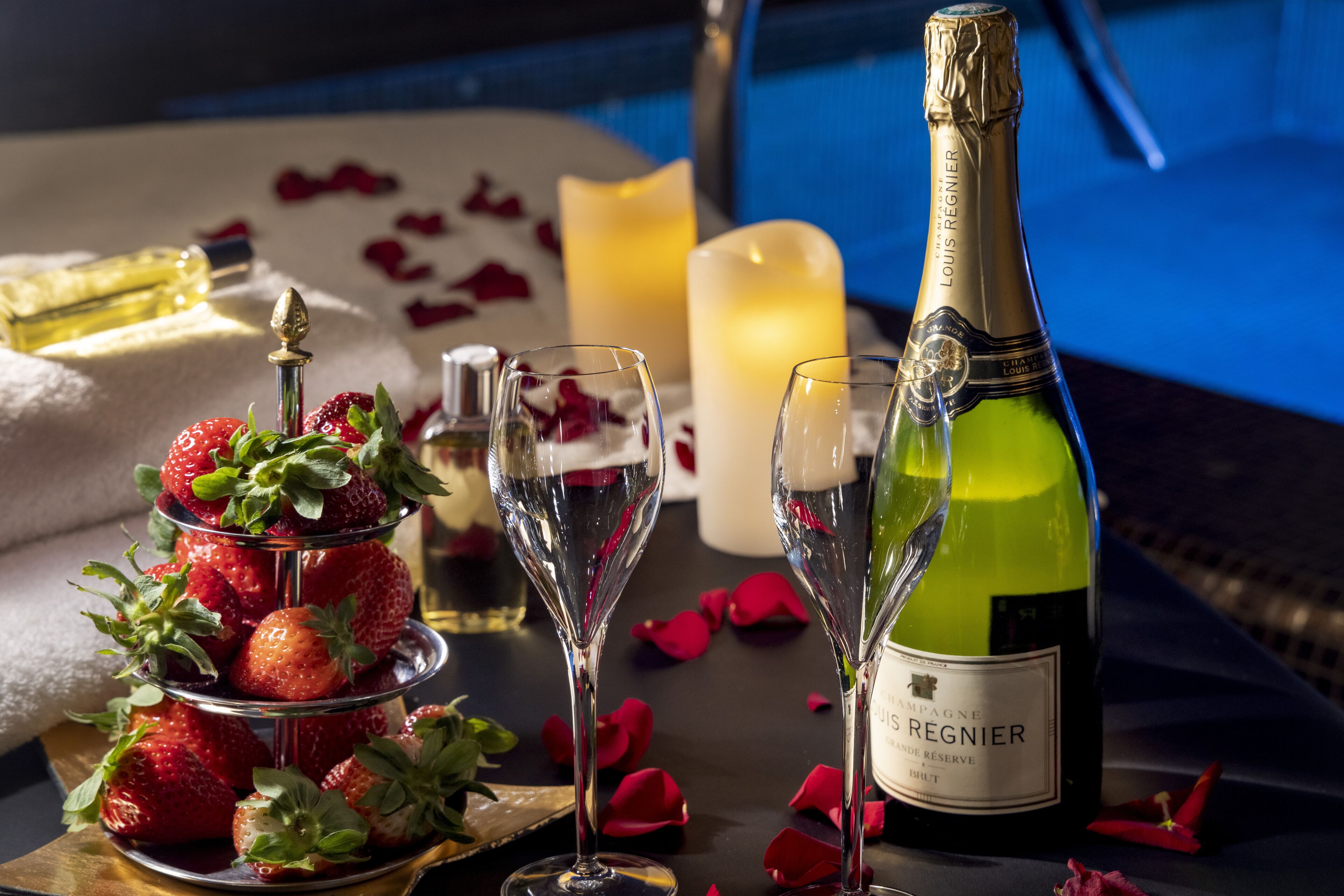 Een tafel met een champagnefles, twee glazen, een schaal aardbeien, kaarsen en rozenblaadjes.