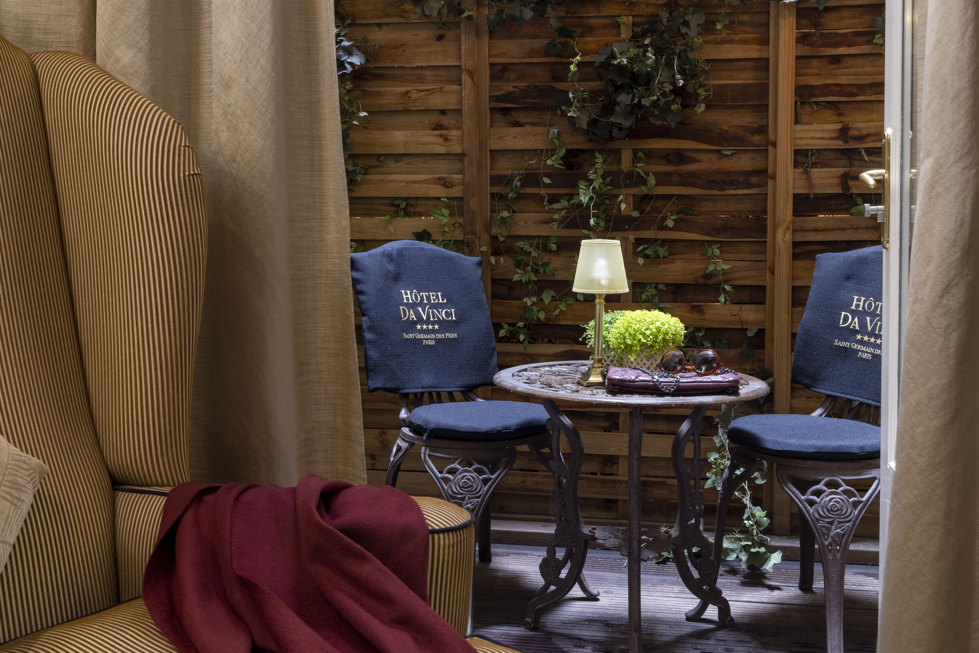 A cozy room with a striped sofa, two chairs with 'Hôtel da Vinci' towels, a small round table with a lamp, and a wooden wall with greenery.