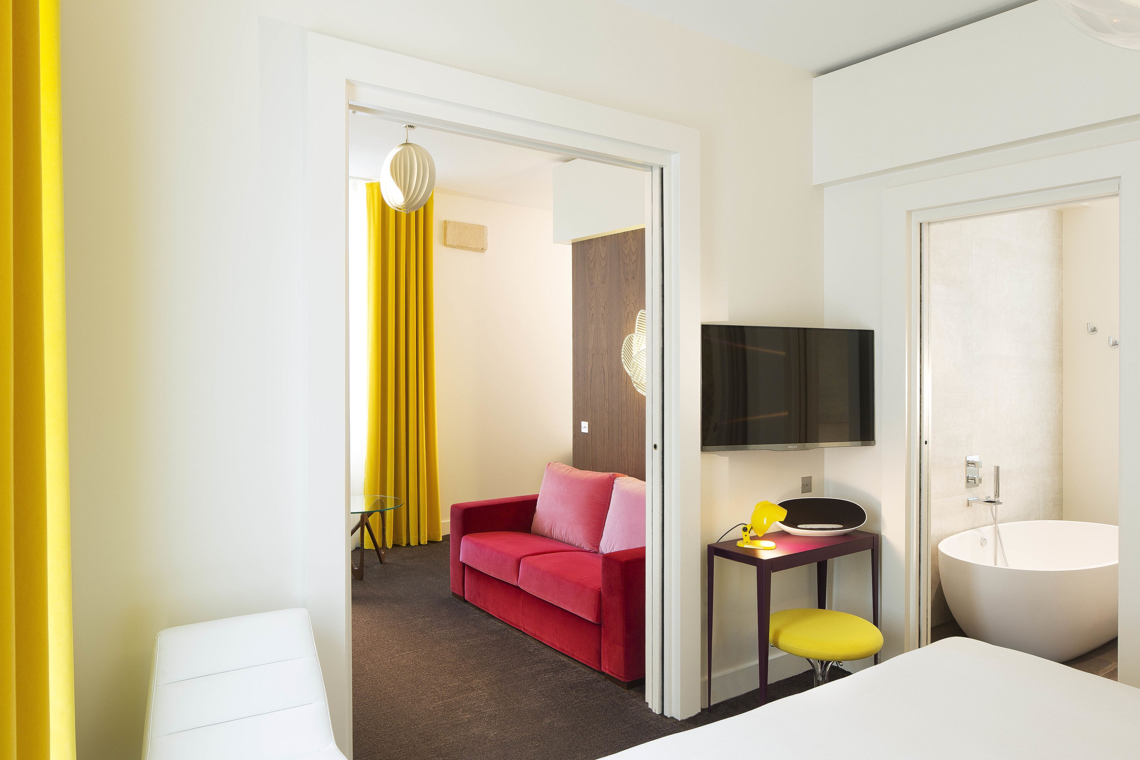 A hotel room with a red sofa, a small table with a lamp, a TV mounted on the wall, and a large bathtub.