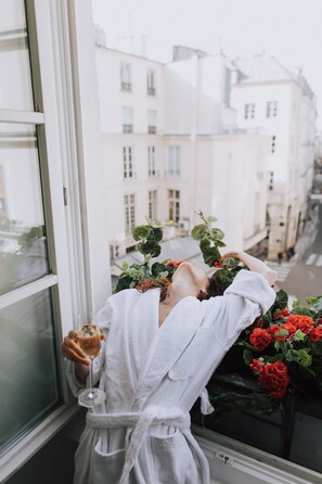 Una persona con bata blanca apoyada en una ventana, sosteniendo una copa de vino y observando una calle con edificios y una planta con flores rojas.