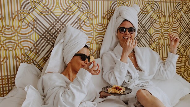 Two people in white robes on a bed with a patterned wallpaper background. - Hôtel Dupond Smith (Paris)