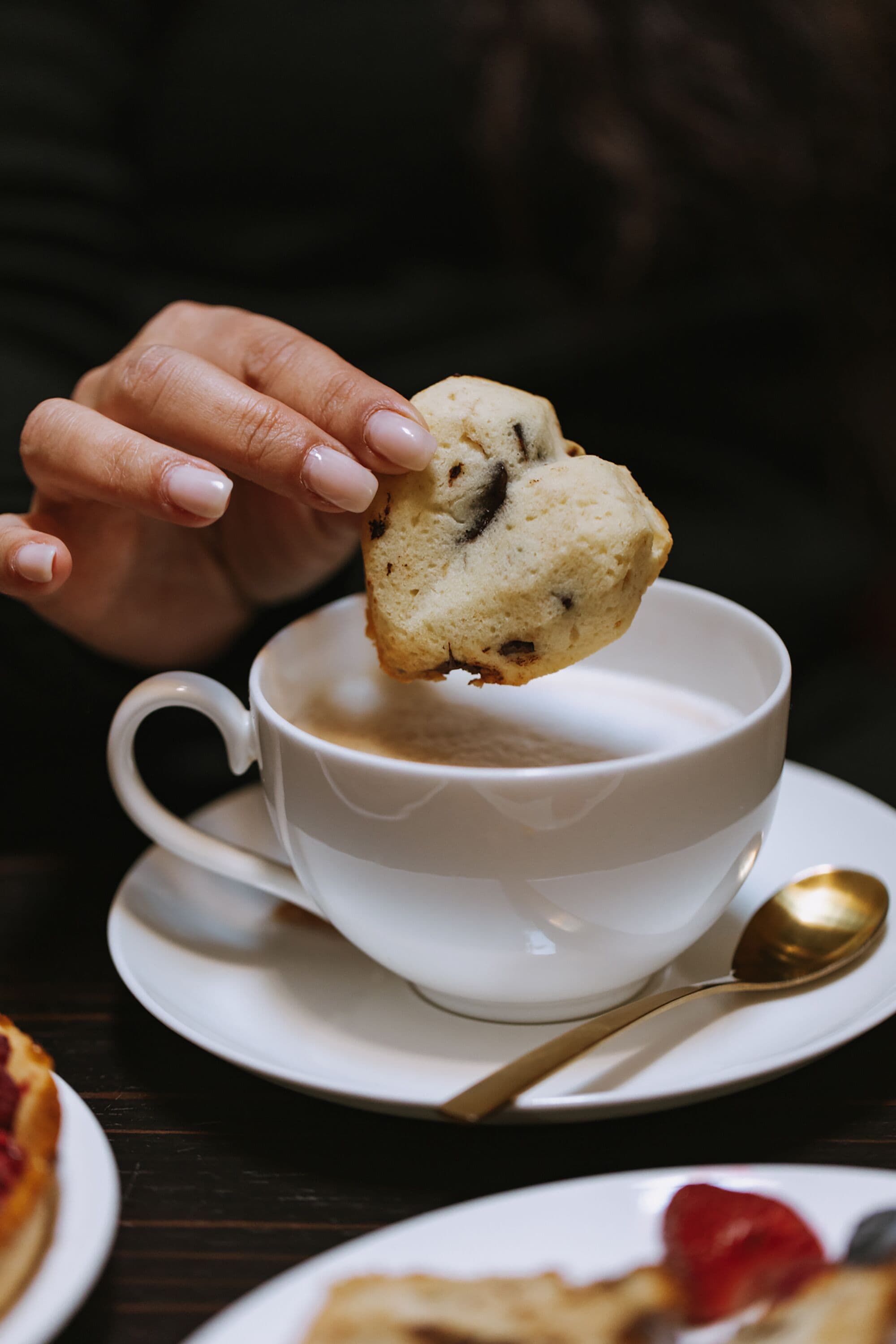 A hand holding a muffin over a cup of coffee.