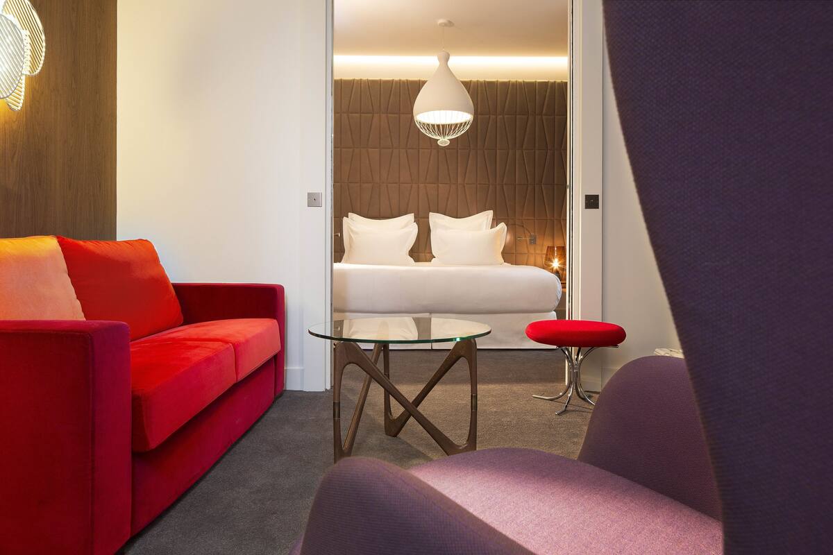 Une chambre d’hôtel moderne avec un canapé rouge, une table en verre, un fauteuil violet, un lit avec du linge de maison blanc et un mur au revêtement original.