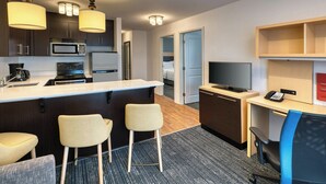 A compact hotel room with a kitchenette, a dining area with two stools, a desk with a computer and phone, and a TV. - Towneplace Suites by Marriott Red Deer (Red Deer)