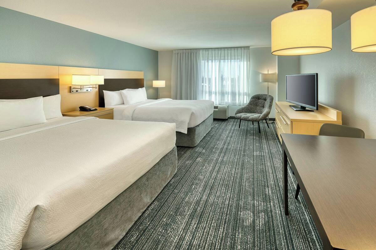 Une chambre d’hôtel avec deux lits, un bureau, une chaise, une télévision et une fenêtre avec des rideaux.