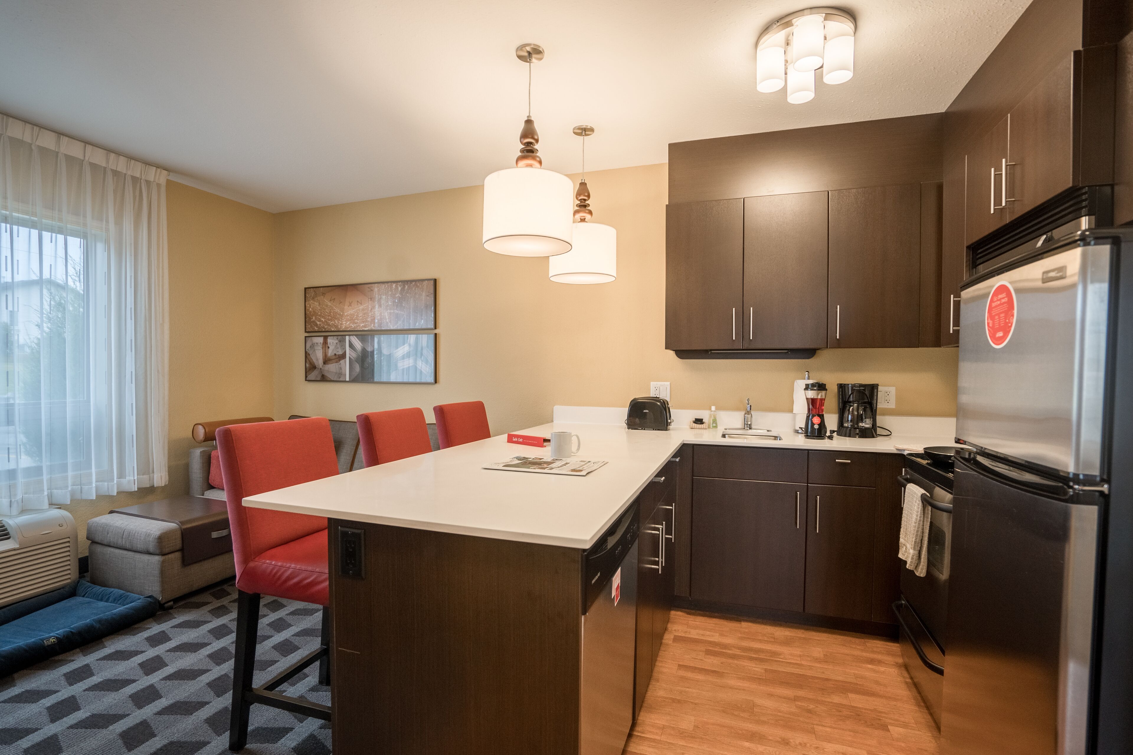 A compact kitchen with a central island, dark wooden cabinets, stainless steel appliances, and a seating area with red chairs.