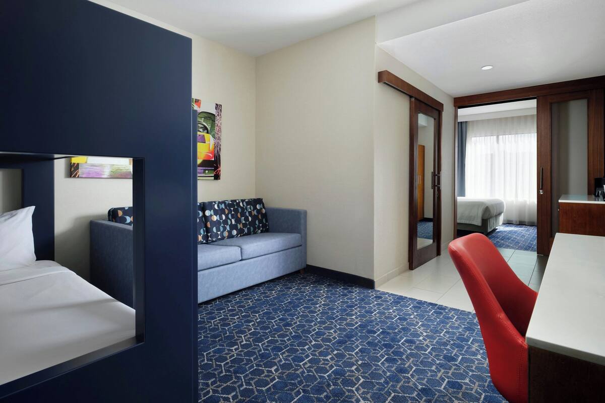 A hotel room with a bed, a blue sofa, a red chair, a small table, and a window with curtains.
