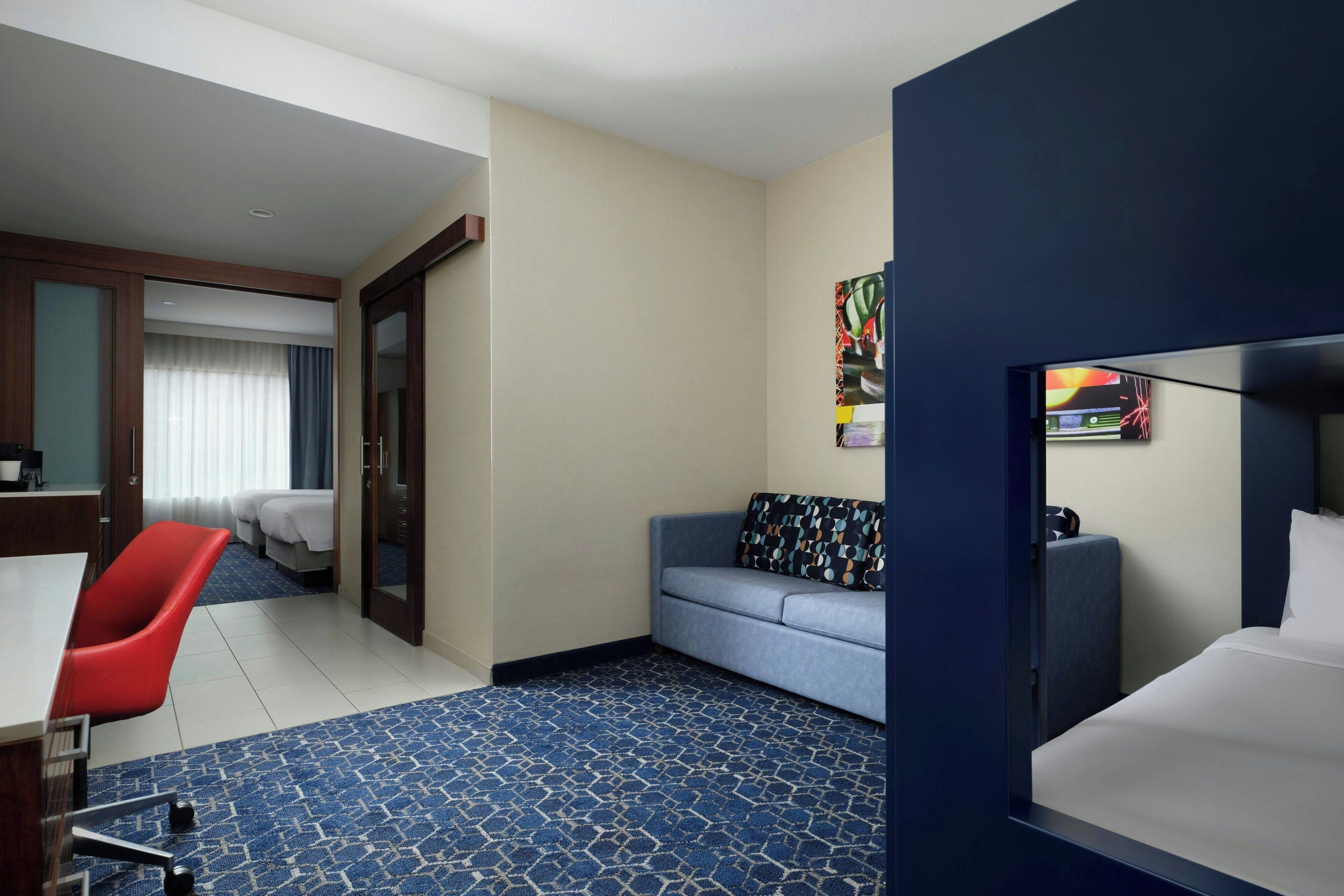 Ein Hotelzimmer mit einem Bett, einem Schreibtisch, einem roten Sessel, einem blauen Sofa und einem farbenfrohen Bild an der Wand.