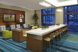 A modern hotel lobby with a long reception desk, comfortable seating, a large TV screen, and large windows offering a view of the city.