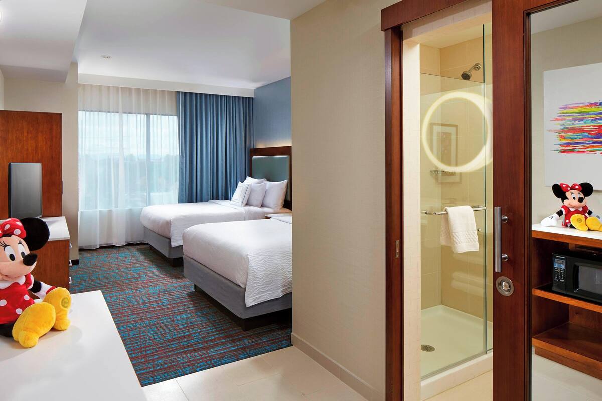 A hotel room with two beds, a shower, a Mickey Mouse plush toy, and a glass-enclosed bathroom with a vanity.