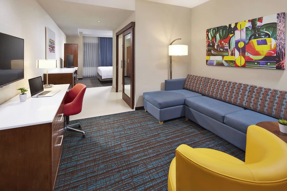 A hotel room with a desk, a laptop, a red chair, a blue sofa, a yellow armchair, a large painting, and a bed with a blue headboard.