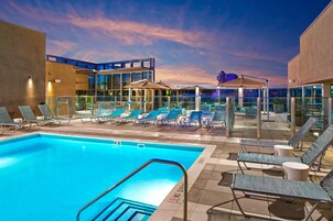 Un área de piscina en la azotea con sillones reclinables y sombrillas, rodeada de muros de vidrio y arquitectura moderna.