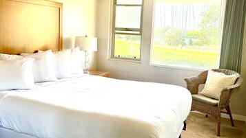 Une chambre d’hôtel avec un grand lit, un ventilateur de plafond, une fenêtre donnant sur les arbres et un fauteuil en osier.