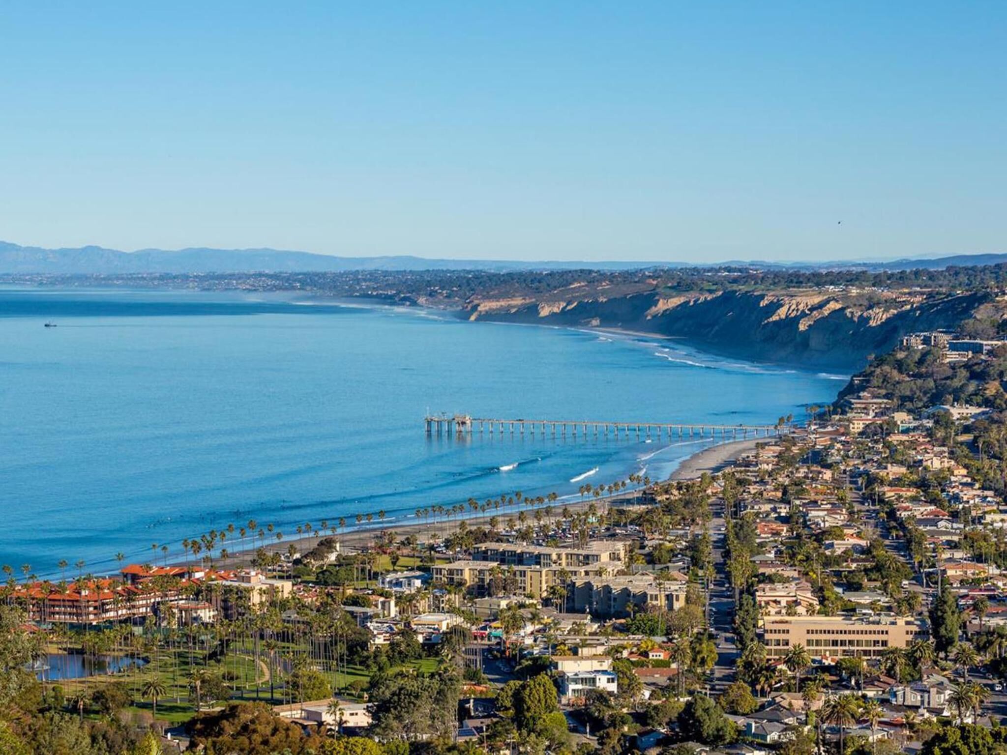 A coastal town with a pier extending into the ocean, residential buildings, and a clear sky.