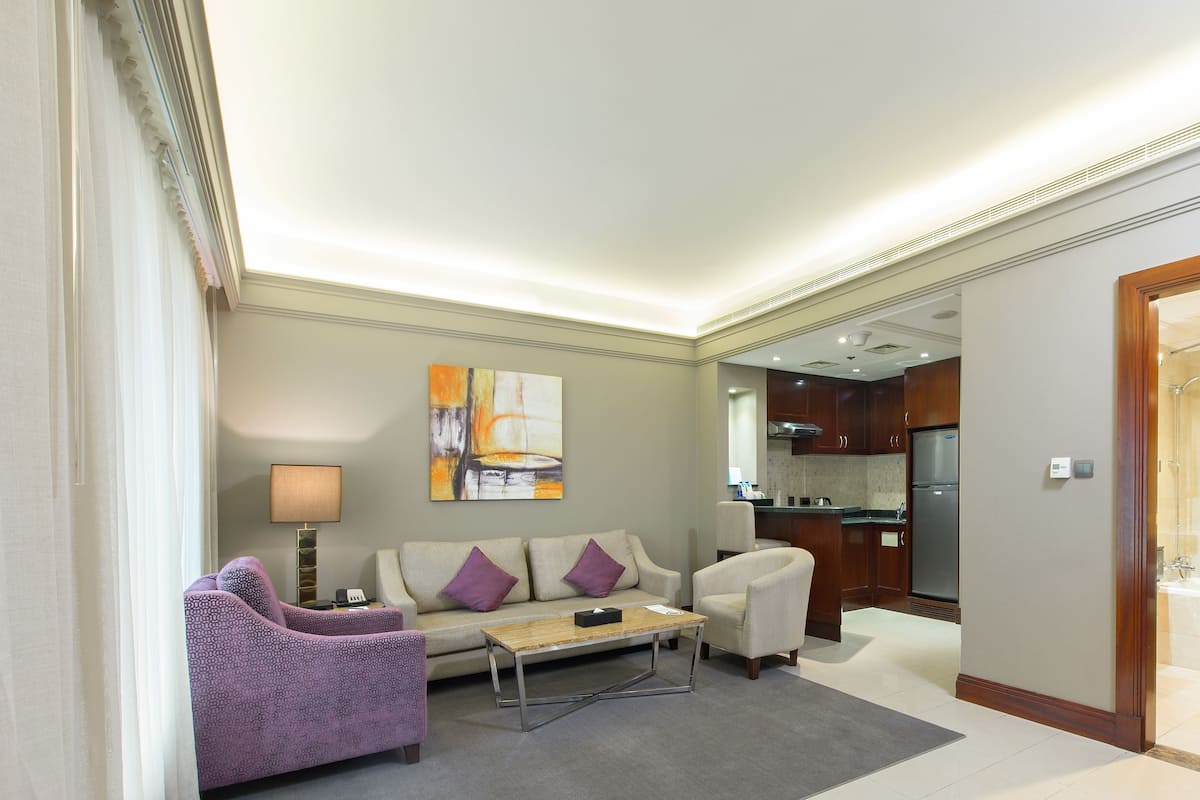 Ein modernes Wohnzimmer mit einer Couch, Sesseln und einem Couchtisch. Im Hintergrund ist eine Küche mit Kühlschrank und Schränken zu sehen.