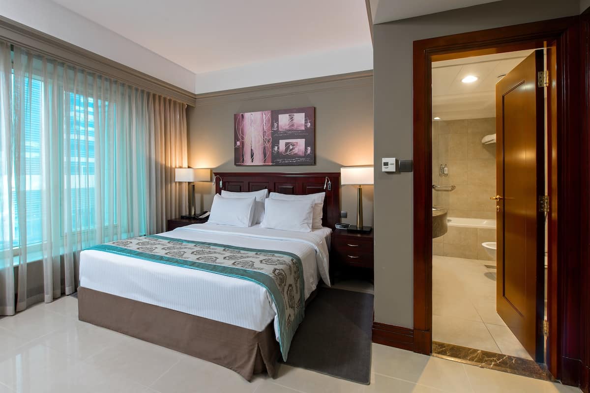 Ein Hotelzimmer mit einem großen Bett, zwei Nachttischlampen, einem Nachttisch, einem Badezimmer mit einer Badewanne und einer Tür zu einem weiteren Zimmer.