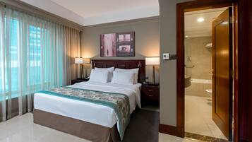 A hotel room with a large bed, two bedside lamps, a nightstand, a bathroom with a bathtub, and a door leading to another room.