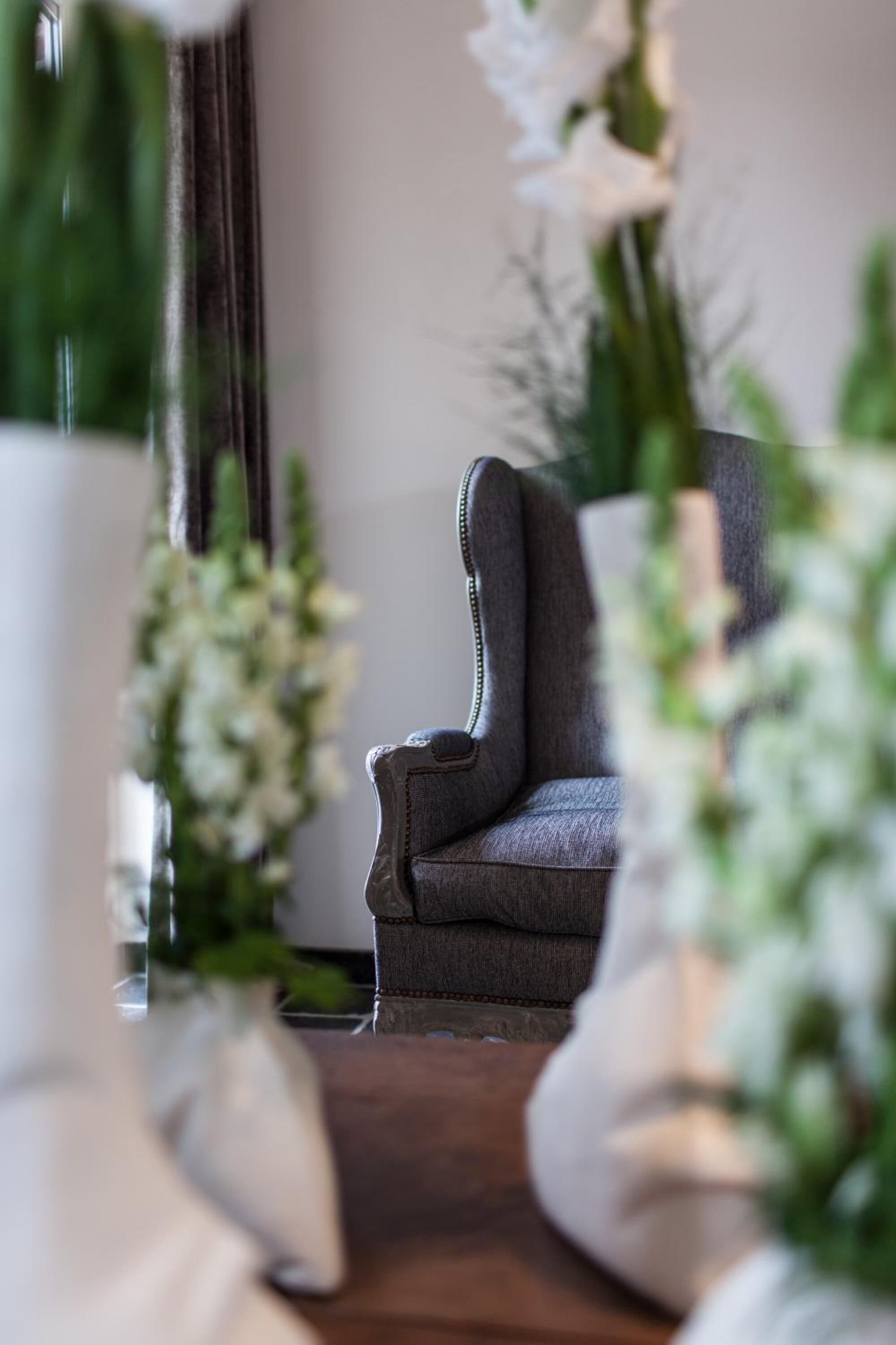 A small, dark-colored armchair in a room with a wooden floor, surrounded by white flowers and greenery.