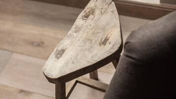 Un tabouret en bois rustique, dont l’assise est usée, est placé sur un plancher en bois, près d’une fenêtre où se trouve une plante en pot.