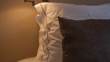 A neatly made bed with white linens and a dark gray pillow. A wall-mounted lamp provides soft lighting.