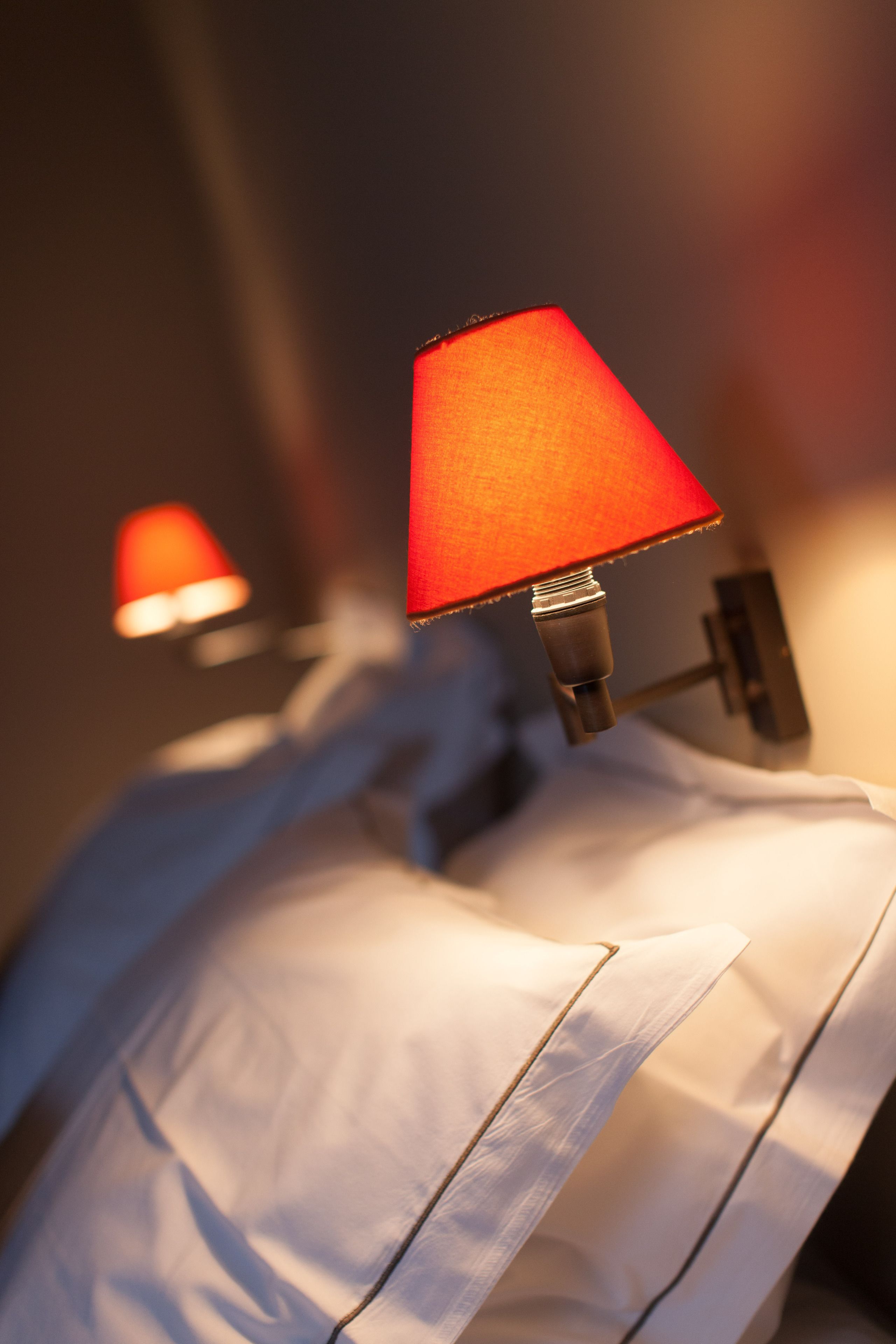 A close-up of a bed with a white pillow and a red lamp on a bedside table.
