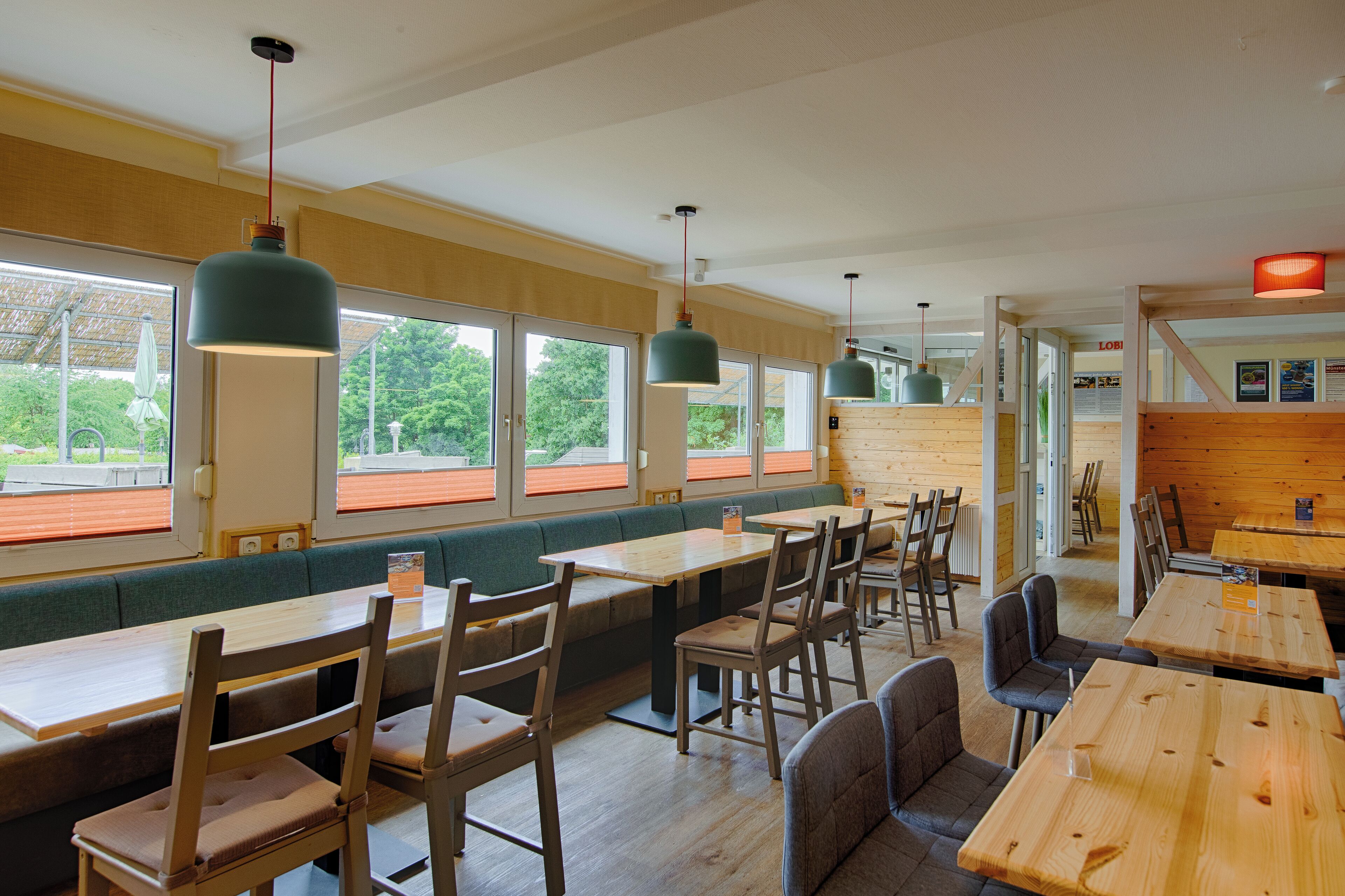 A restaurant with wooden tables and benches, large windows with orange blinds, and hanging green pendant lights.