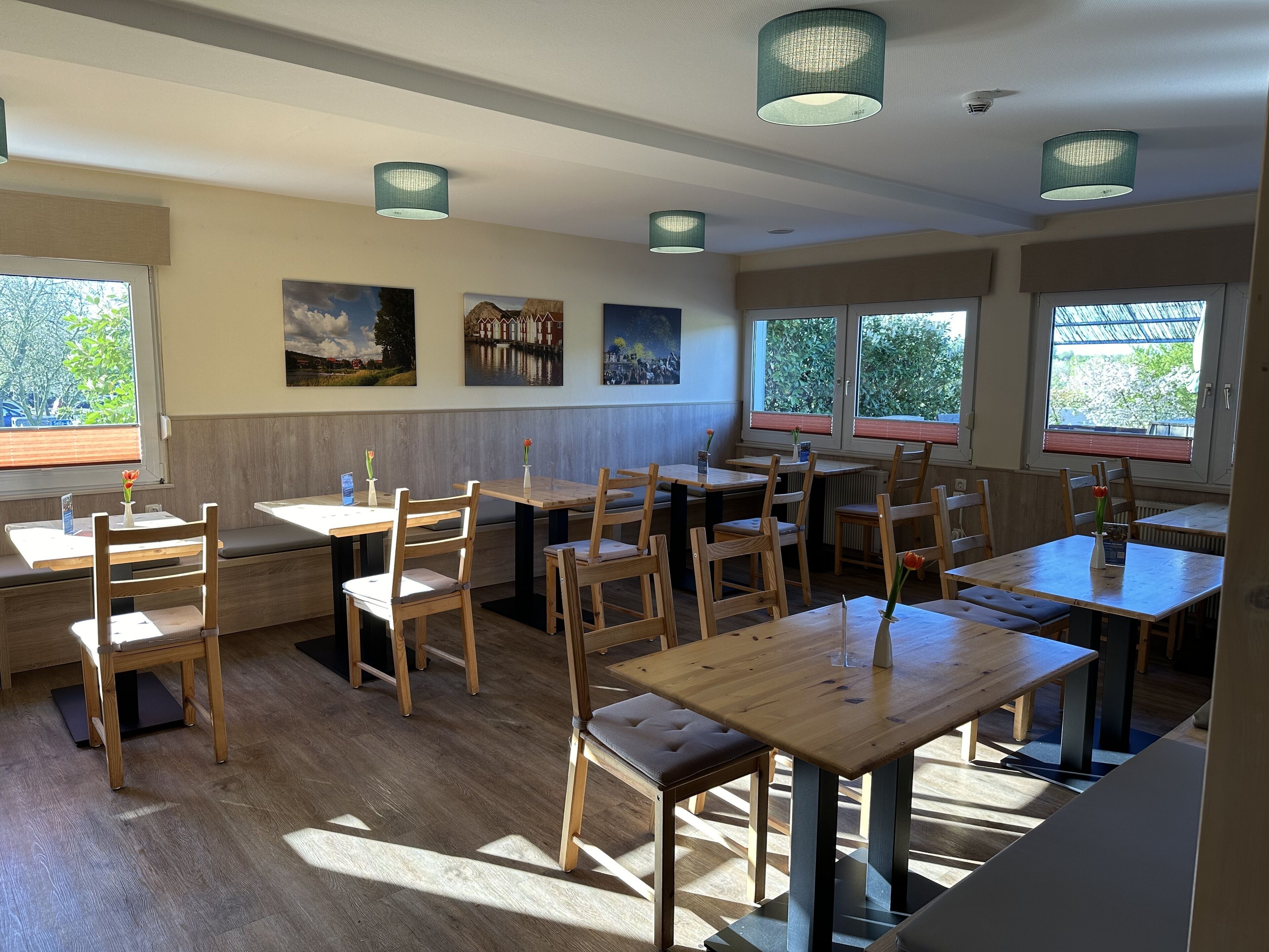 A restaurant with wooden tables and chairs, large windows with blinds, and artwork on the walls.