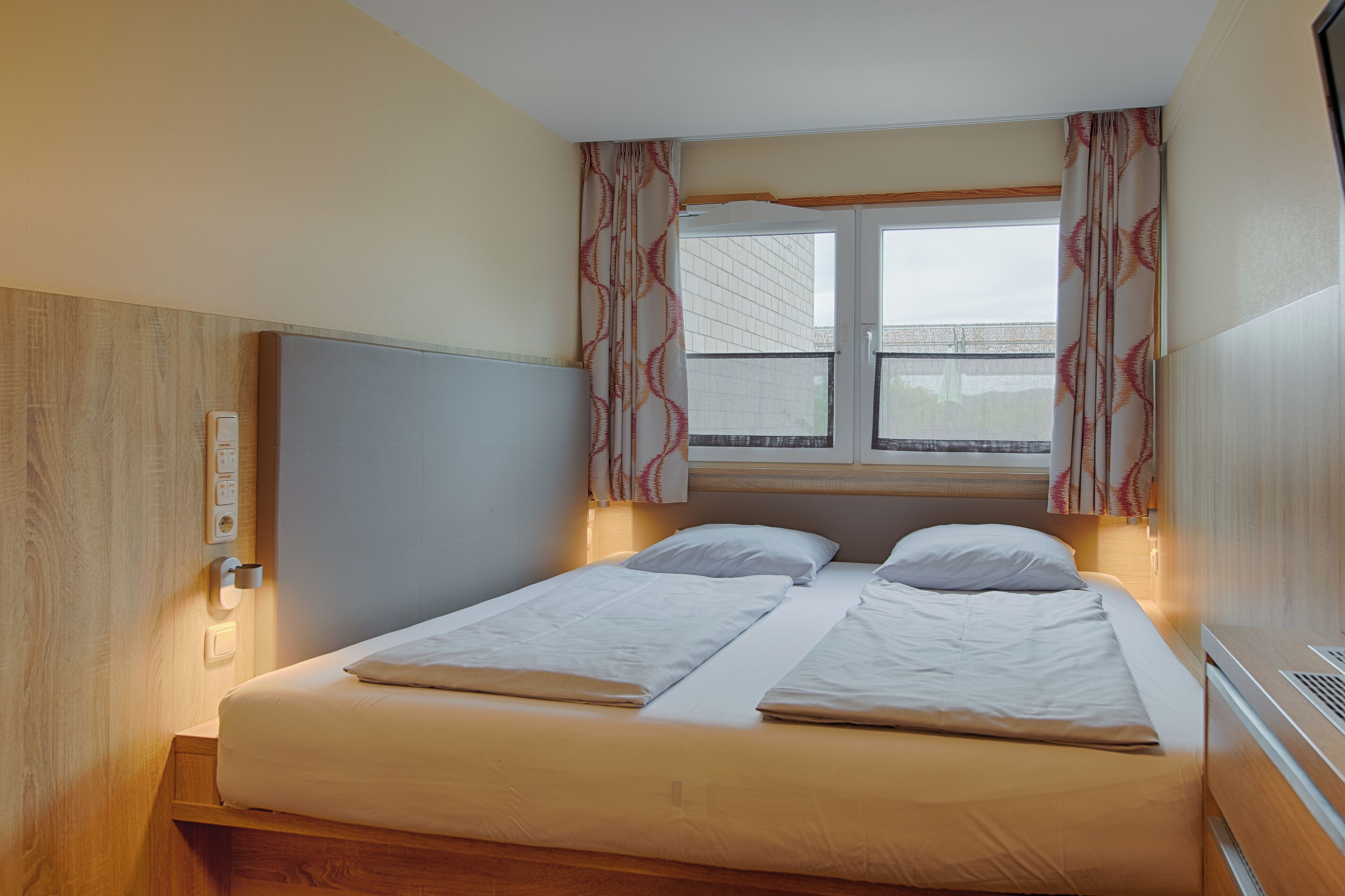 A double bed with white bedding, a wooden headboard, and a window with patterned curtains.