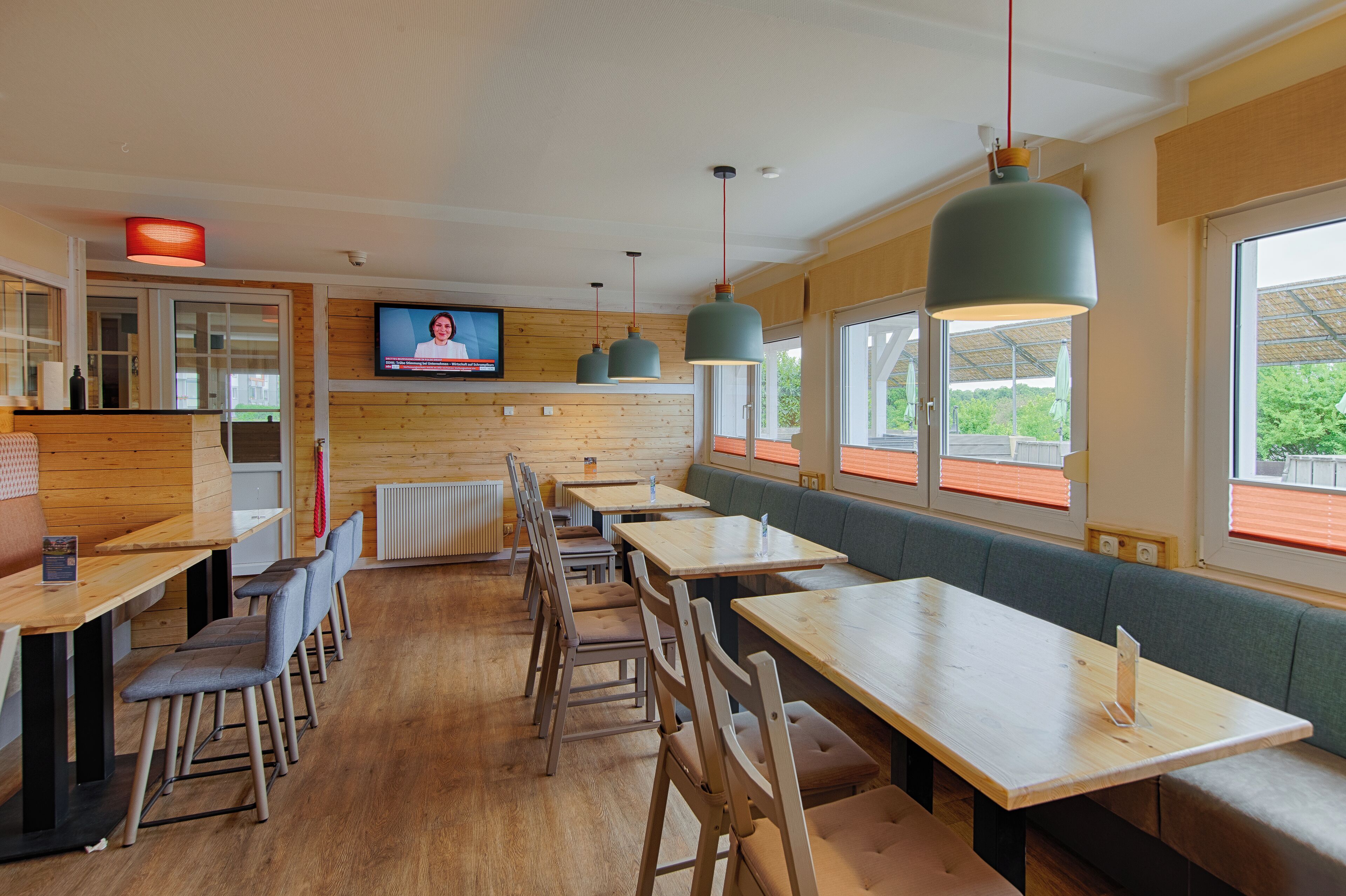 A restaurant with wooden walls, multiple tables and chairs, a television screen, and large windows with a view of greenery.