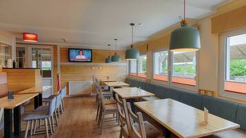 A restaurant with wooden walls, multiple tables and chairs, a television screen, and large windows with a view of greenery.